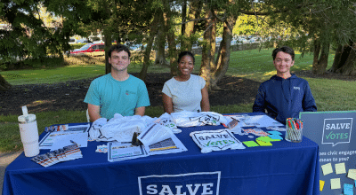 Three SalveVotes student members sit behind a voter engagement table outdoors on campus.