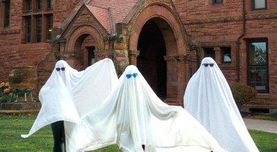 Three students dressed as ghosts in front of red building.