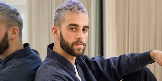 Young bearded man with short purple hair leaning against a mirror