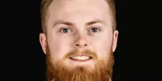 Male student with reddish hair and beard, in white shirt and dark solid color tie looking at camera