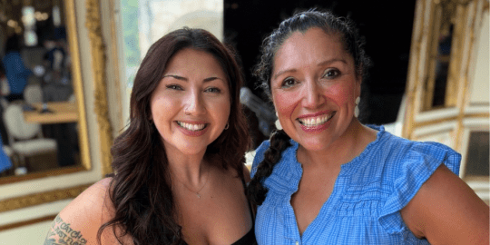 Acevedo (left) in a black top and Jennifer De Leon in a blue dress together and smiling at the camera.