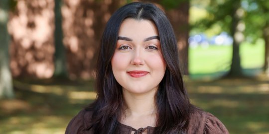 Young alumna, brown hair, brown shirt, standing outside smiling