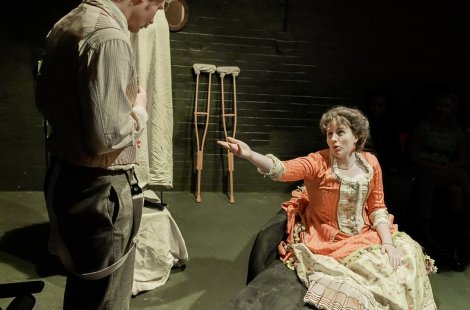 Actress in period costume gesturing to a man, crutches in the background