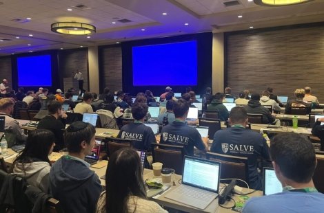 Students at tables in an Excel competition, facing big screens