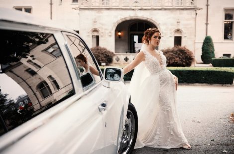 A woman in a white wedding dress leans on a white limo in front of a mansion.