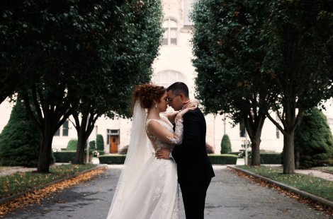 A bride and groom embrace along a tree-lined driveway leading to an elegant mansion.