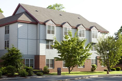 Three-story residential building with light siding, brick lower walls and trees in front.