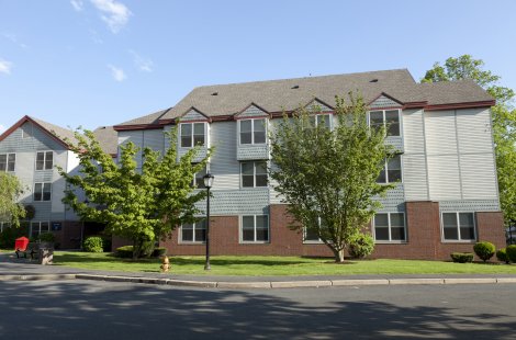 Three-story residential building with light siding, brick lower walls and trees in front.