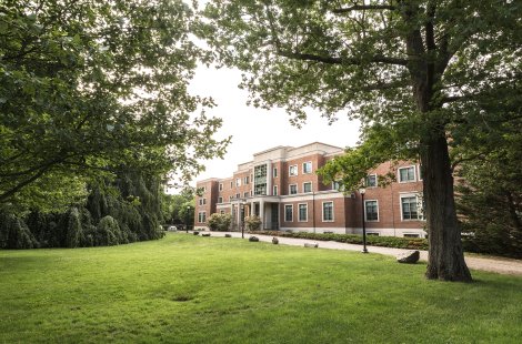 A brick building with large windows and a columned entrance, surrounded by a green lawn and tall leafy trees.