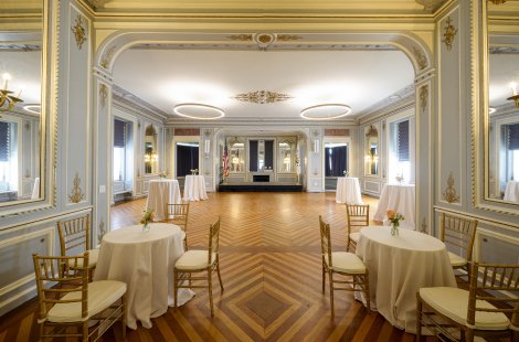 A large ballroom with ornate walls, set with small cocktail tables and chairs.