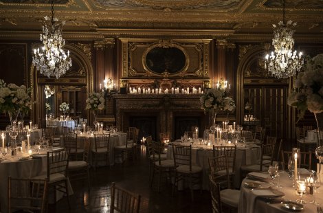 An elegant ballroom with ornate gold detailing, crystal chandeliers and round tables set for a formal wedding reception.