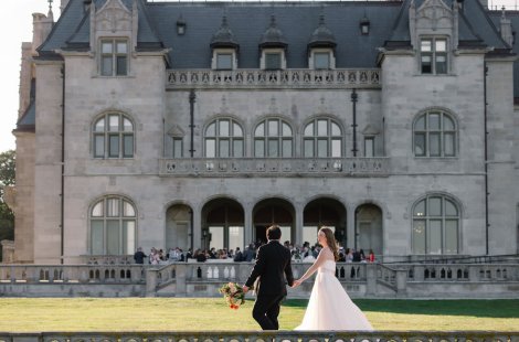 A couple in wedding attire walks hand in hand across a lawn in front of a large historic stone mansion.
