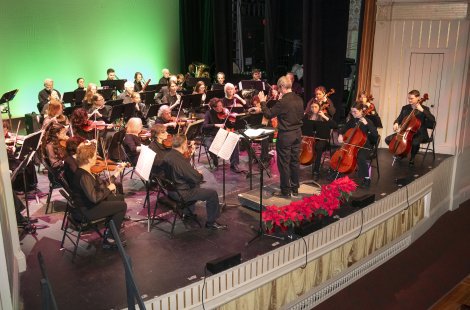 Orchestra performing on a theater stage, with a conductor leading seated string, woodwind, brass and percussion musicians dressed in black.