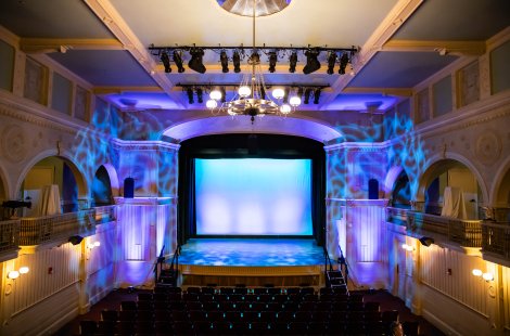 An ornate theater auditorium with balcony seating and a lit stage featuring a blue backdrop and overhead stage lights.