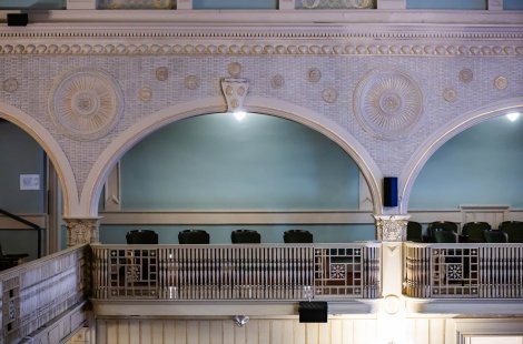A decorative balcony inside a historic theater with arched openings, carved wall details and rows of seats.