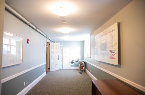 Long hallway with gray walls, white trim, ceiling lights, exposed piping and doors at the far end.