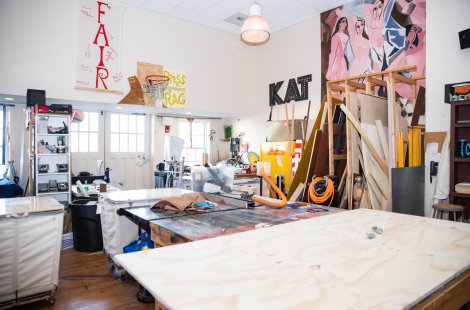 Workshop with large worktables, tools, wood panels, shelves and colorful artwork on the walls.