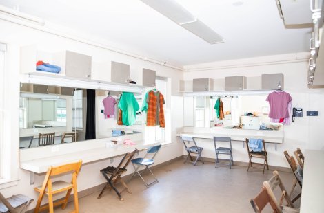 Dressing room with mirrors, countertop vanities, folding chairs, overhead cabinets and garments hanging on racks.