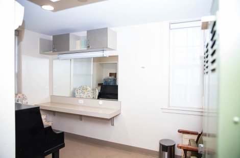 Accessible dressing room with long counter, mirror, overhead cabinets, window with shade and a piano partially visible at left.