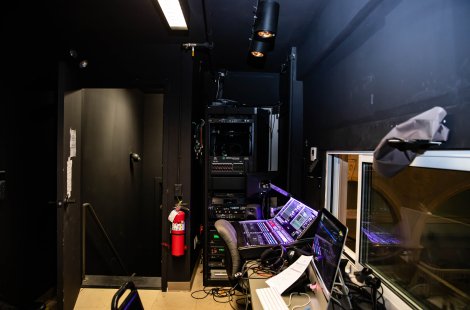 Audio control booth with mixing console, computer monitors, equipment racks and a window overlooking a theater.