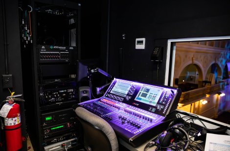 Close-up of audio mixing console and rack-mounted equipment in a theater control booth, with stage visible through a window.