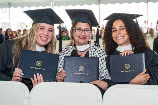 Three graduate students sitting and smiling with their diplomas in hand, in their caps and gowns.