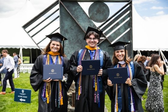 Three students smiling and holding their diplomas in their cap and gowns outside.
