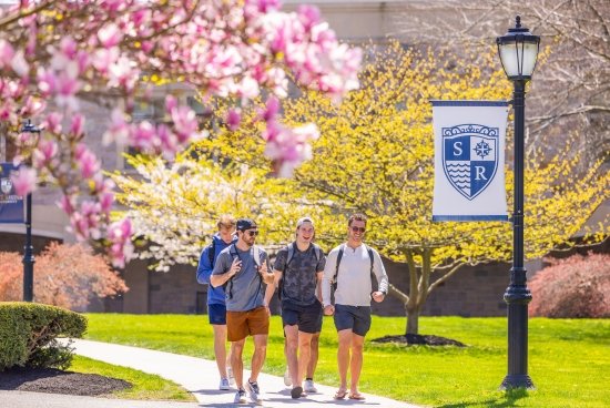 Four mail students walking on campus by tree in full pink bloom, near light with salve crest banner