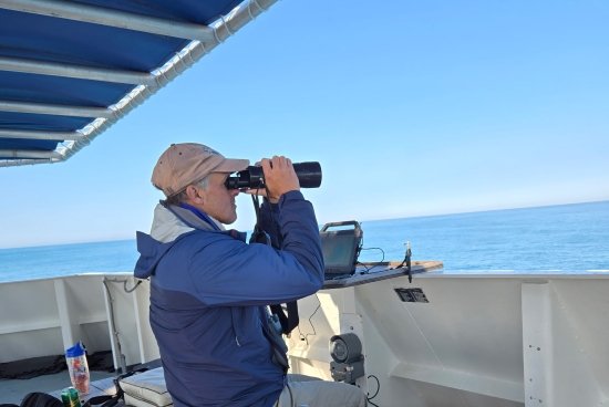Chace on a boat looking out into the water with binoculars.