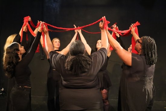 Performers in a circle with arms in the air, holding a red ribbon.