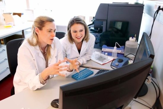Jillian Wise and another collegue sitting in white lab coats discussing something they are viewing on the computer in front of them.