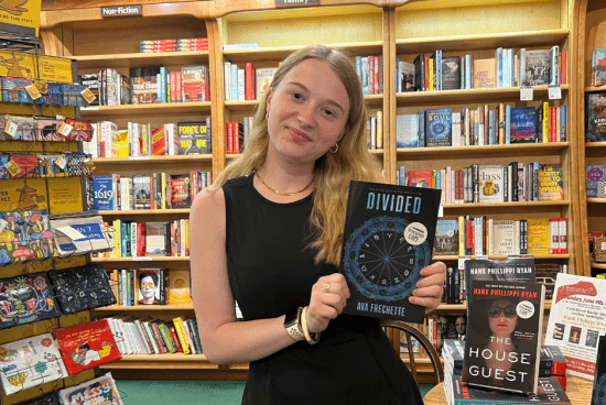 Ava wearing a black dress in a bookstore holding a copy of her first book "Divided"