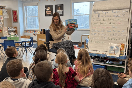 Teacher reads a picture book to students seated on a classroom rug.