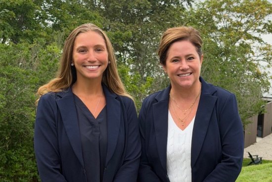 Two women stand together outdoors with Newport Harbor in the background.