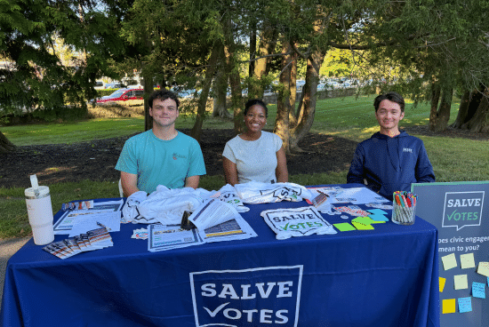 Three SalveVotes student members sit behind a voter engagement table outdoors on campus.