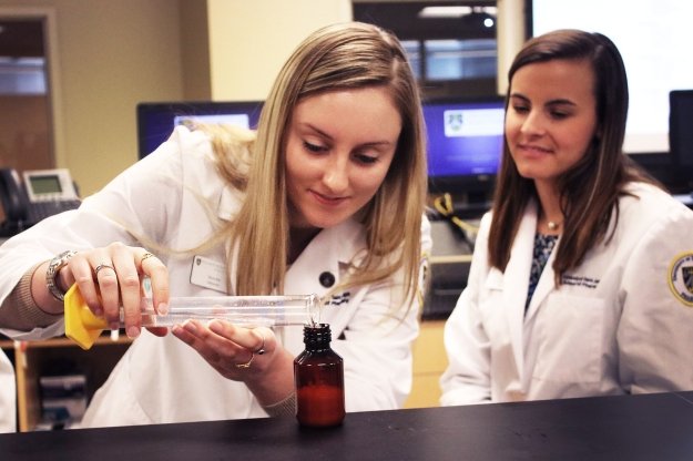two students in lab coats pouring liquid from beaker