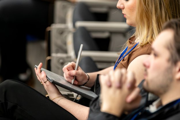 A person seated in a conference setting writes notes on a tablet while another attendee sits nearby out of focus.