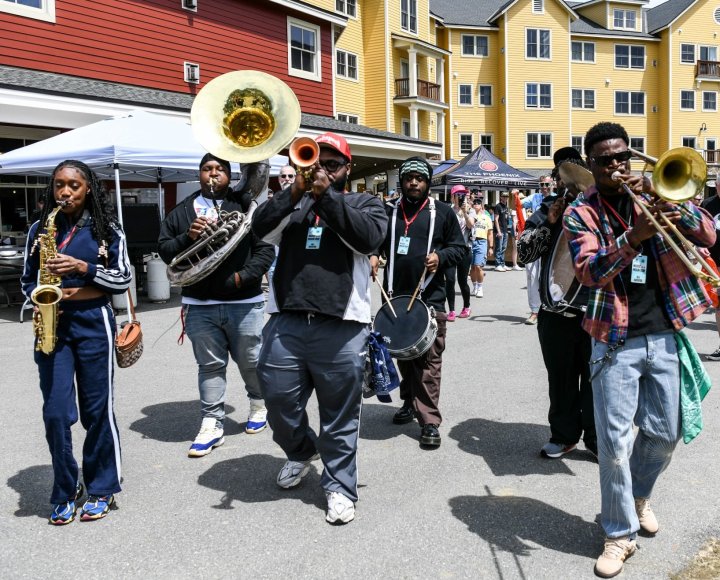 A marching brass band performs outdoors, with musicians playing saxophone, trumpet, trombone, tuba and drums.