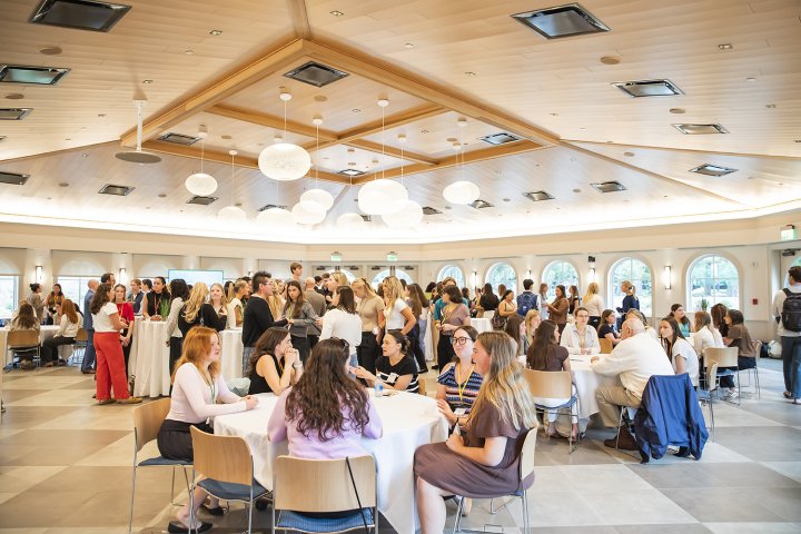 A large, brightly lit event hall with groups of people seated at round tables and standing in conversation during a networking event.