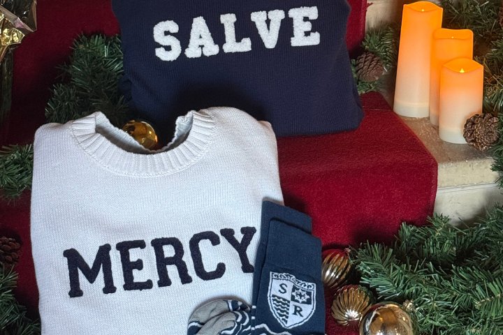 Folded navy and white sweaters embroidered with "Salve" and "Mercy," displayed with navy socks and Christmas decorations.
