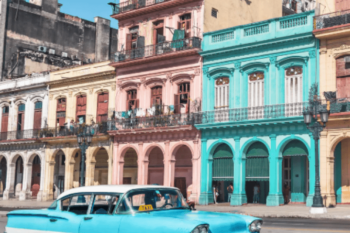 A classic American car drives past a block of pastel buildings