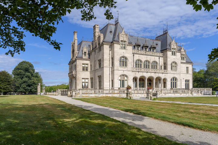 A stone mansion with ornate gables and arched windows, set on landscaped lawns under a blue sky.