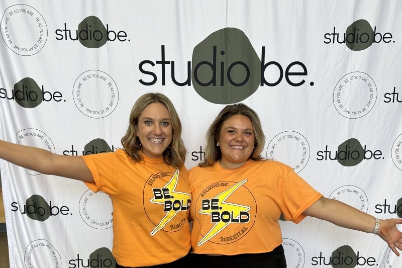 Two girls smiling in matching orange t-shirts in front of a Studio Be backdrop in black and white.
