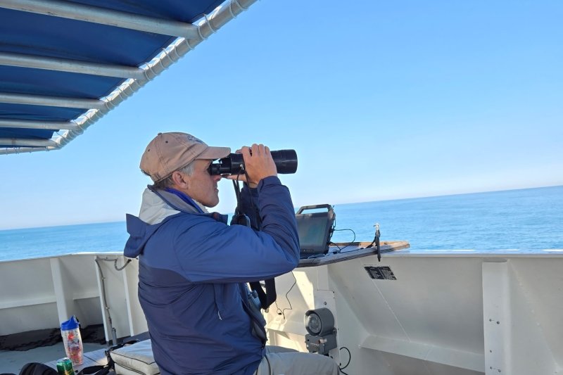Chace on a boat looking out into the water with binoculars.