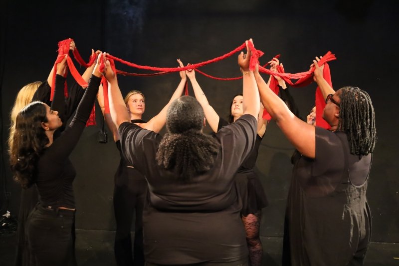 Performers in a circle with arms in the air, holding a red ribbon.