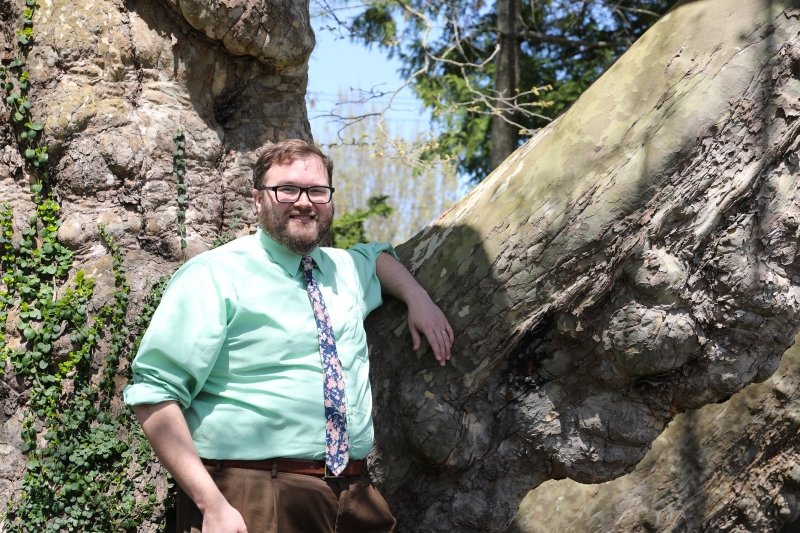 Ben Hohman in an aqua colored button down shirt and pink and blue tie, smiling with his arm rested up against a tree.