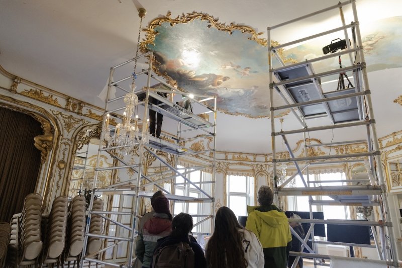 A group of people with their backs turned to the camera looking up at an oval-shaped ceiling painting in Ochre Court's ballroom.