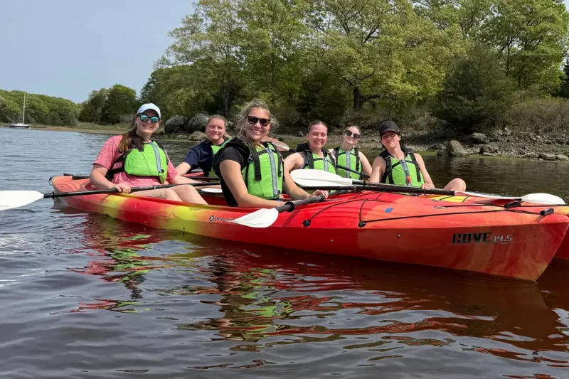 Group of six female students in lime green life vests sitting it two red kayaks on a lake with trees in the background, smiling at camera.