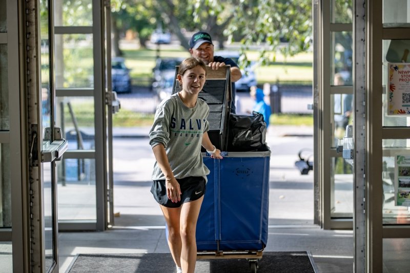 Student in Salve shirt pulling a cart of her stuff behind her as she walks into her residence hall.