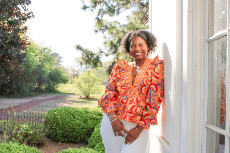 Professional landscape shot of Elodie in colorful orange blouse, smiling while leaning against a white building with trees in background.